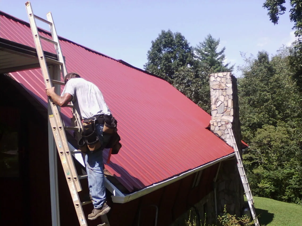 Roofer on a ladder beside a red metal roof and stone chimney — Soffit & Fascia Repair by Roofing Brothers in Rock Hall